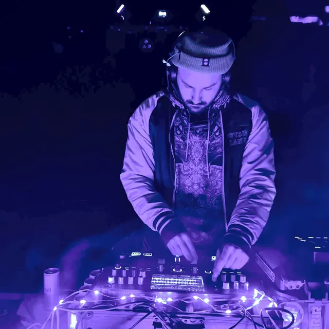 AR/IS at the decks in a dark room lit deep blue, wearing a beanie with headphones round his neck and a satin bomber jacket, both hands on the mixer