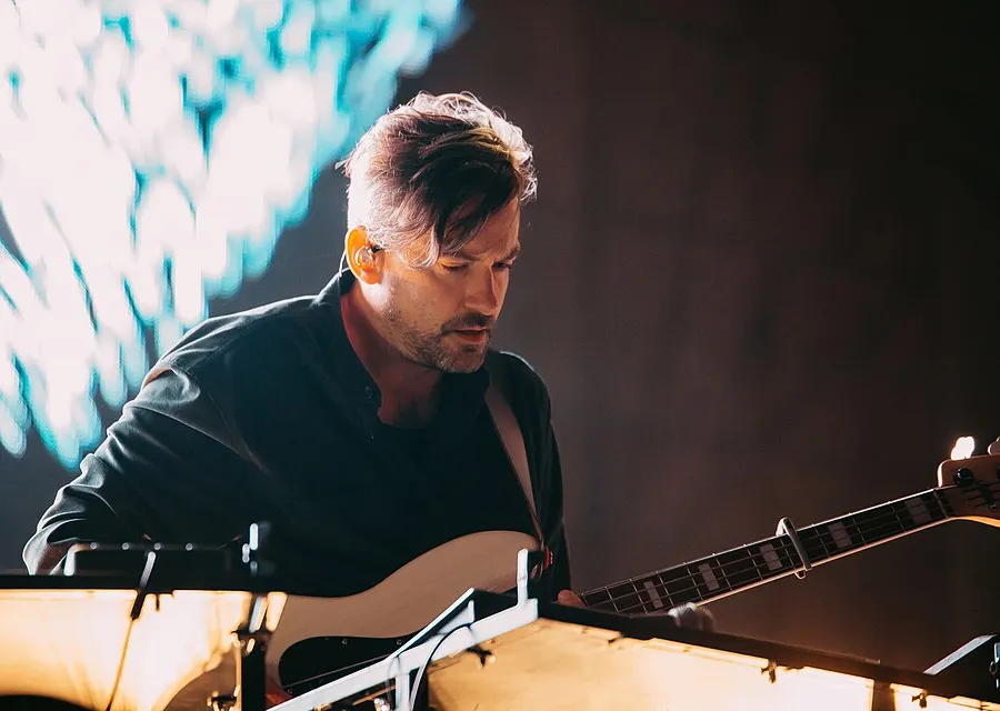 Bonobo playing bass on a dark stage, wearing an in-ear monitor, lit from behind by a pale blue backdrop