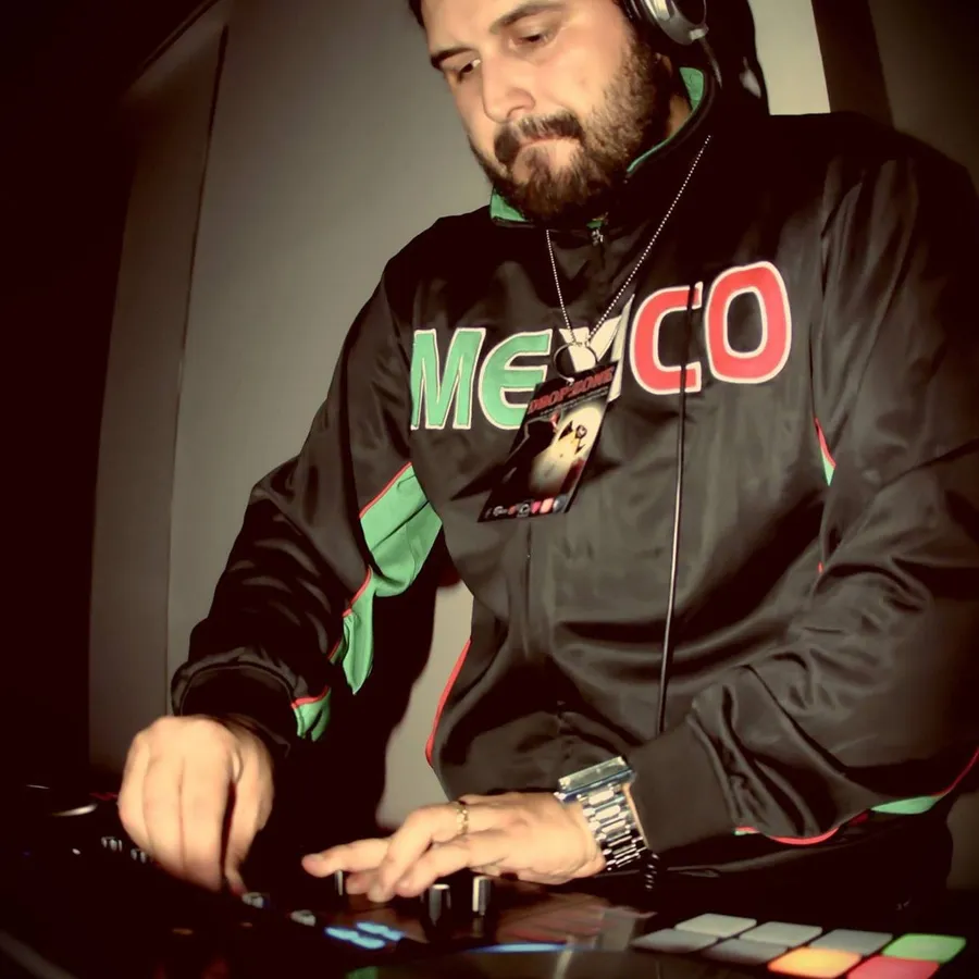 DJMOJO at the controller in headphones, wearing a Mexico track jacket, lit from the side in a dark room