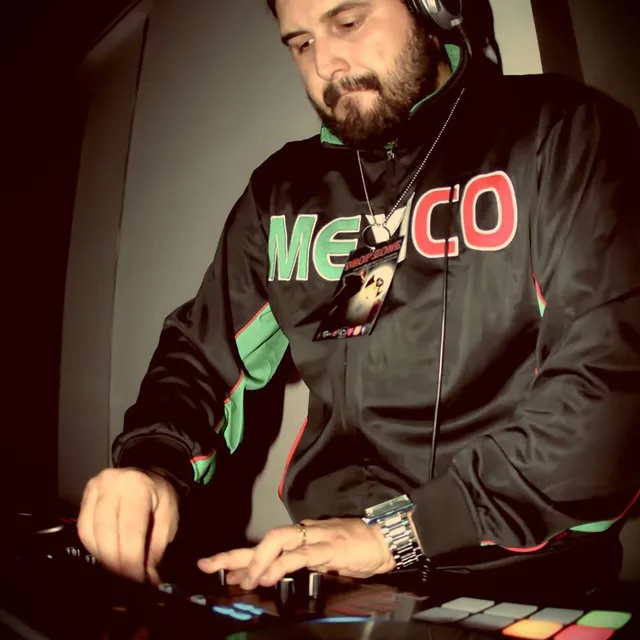 DJMOJO at the controller in headphones, wearing a Mexico track jacket, lit from the side in a dark room