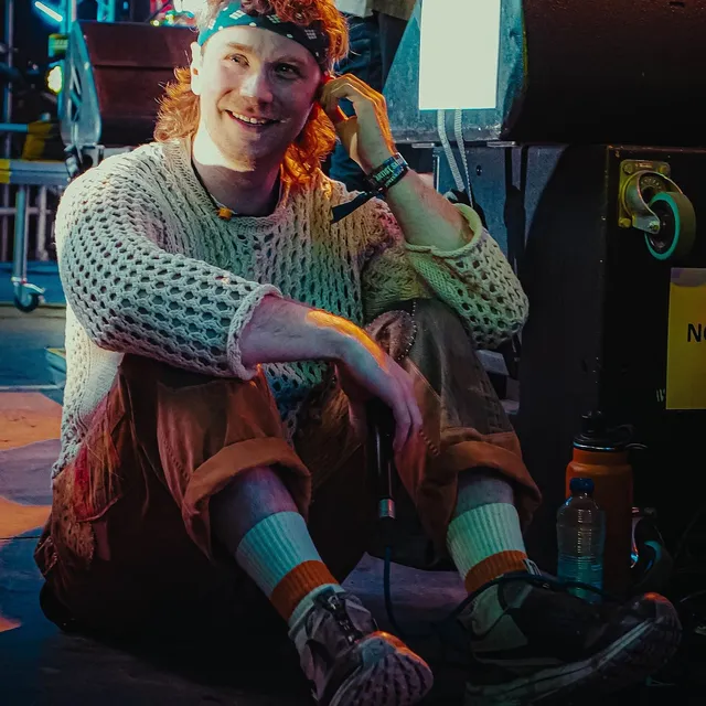 Fox Stevenson sitting on the floor backstage in a loose knitted jumper and a bandana, smiling towards the camera, stage rigging and flight cases behind him