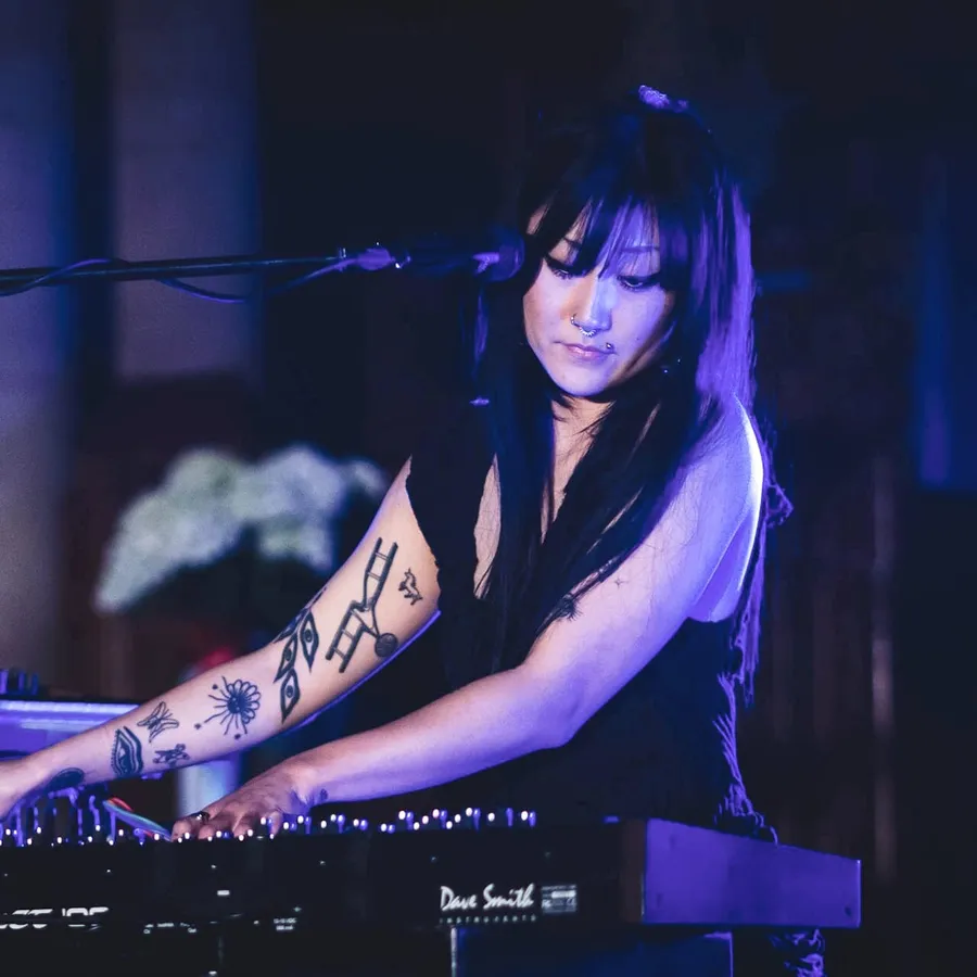 Hinako Omori on stage under blue light, head down and both hands on the keys of a synthesiser, a microphone angled in from the left
