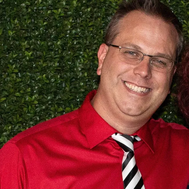 HouseGruv smiling in a red shirt and a black-and-white striped tie, in front of a green hedge wall