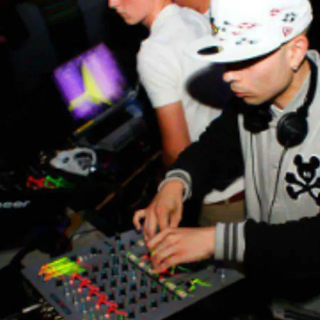 Two DJs side by side at a club mixer in low light, the nearer one in a white cap and headphones, screens glowing behind them
