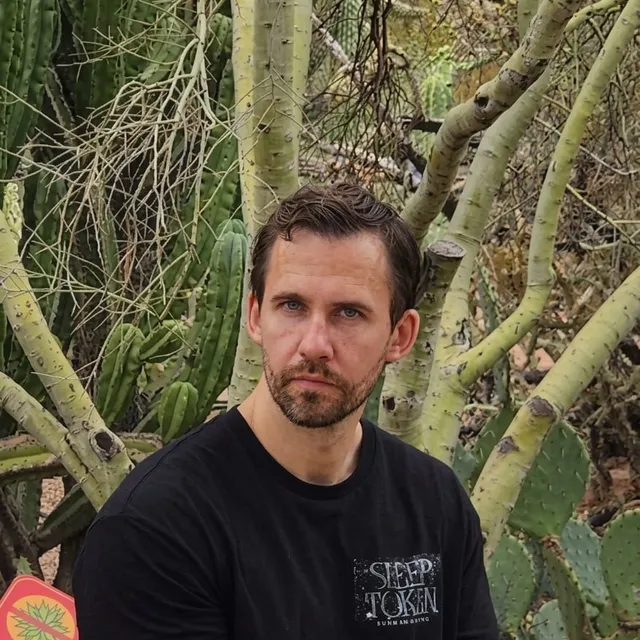 MaDonkey in a black t-shirt in front of tall cacti in an Arizona desert garden, looking straight at the camera