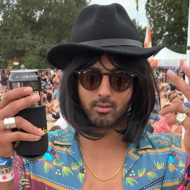 Miami Papi in a wide-brimmed black hat and round sunglasses, an open patterned shirt and a gold chain, holding a drink up towards the camera on a festival field