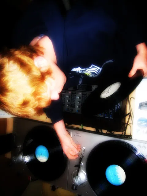 Will See seen from above at the decks, one hand lifting a record and one between two turntables, over a mixer in low blue light